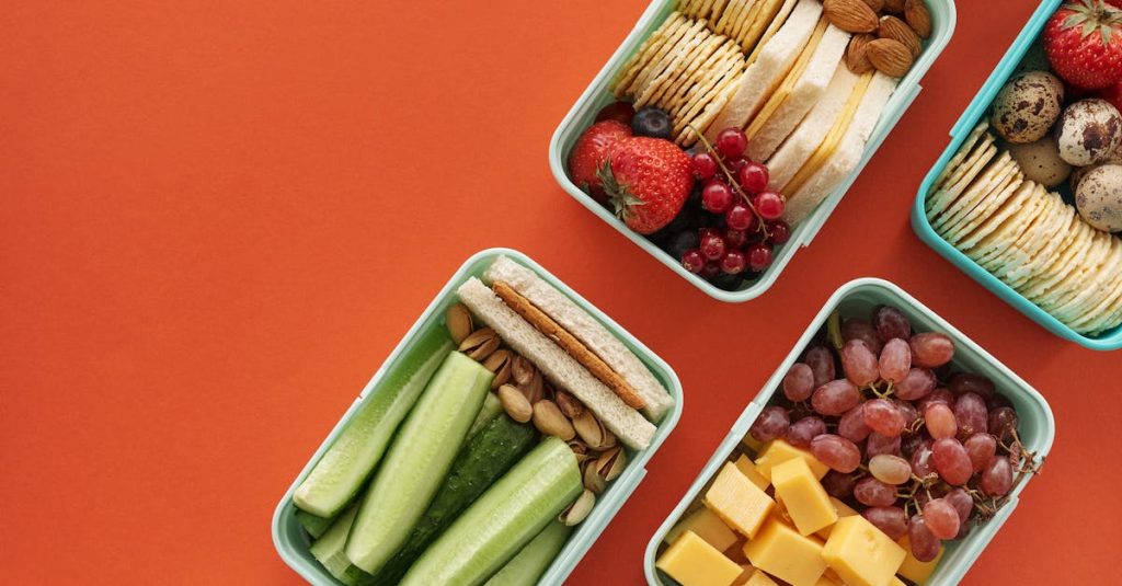 Flat lay of assorted healthy lunchboxes with fruits and nuts against vibrant orange background.
